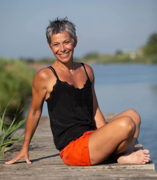 Sophie souriante est habillée d'un short orange et un top noir. Elle est assise sur un ponton gris en bordure de canal.